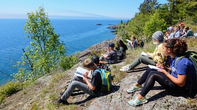 Students sitting on a cliff side, looking at the ocean and making notes in notebooks