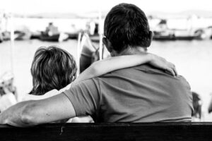 Two people sit on a bench with arms around each other, seen from behind, overlooking a harbor.