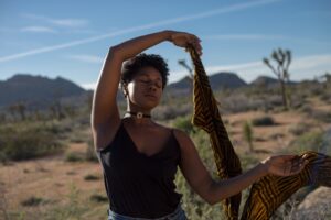 Black person with eyes closed lifting up a gold and black scarf. Desert landscape in background.